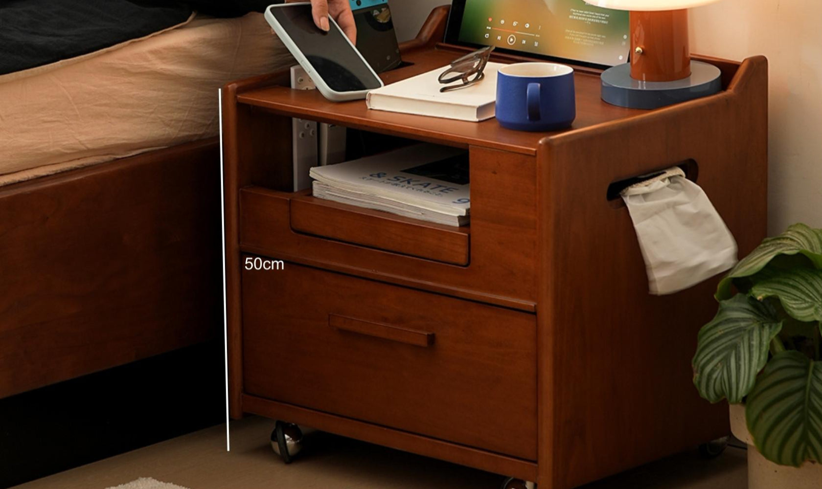 Wooden nightstand with a drawer, books, and a tablet on top next to a bed.