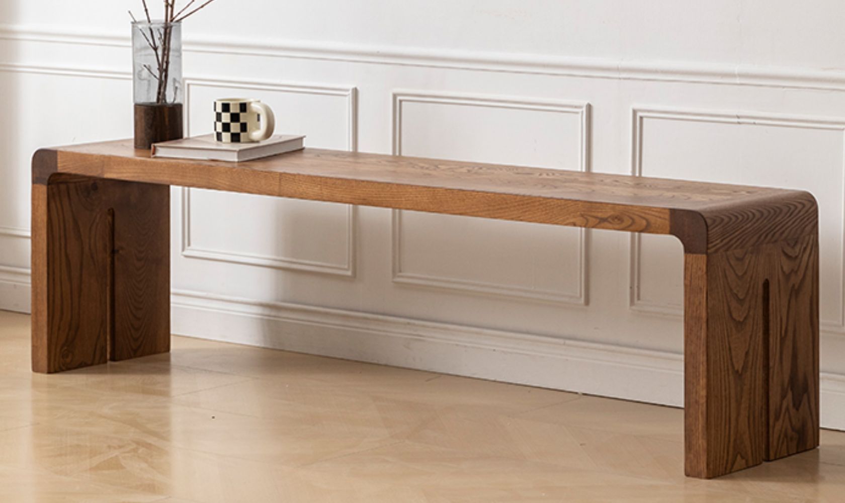Wooden bench with a vase and books on a light wood floor against a white paneled wall.