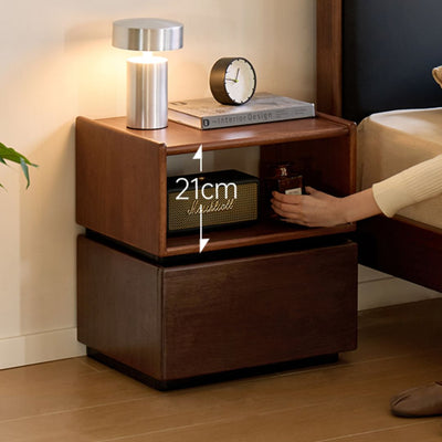 Wooden nightstand with a lamp, clock, and small box on top, with a person reaching for an item.